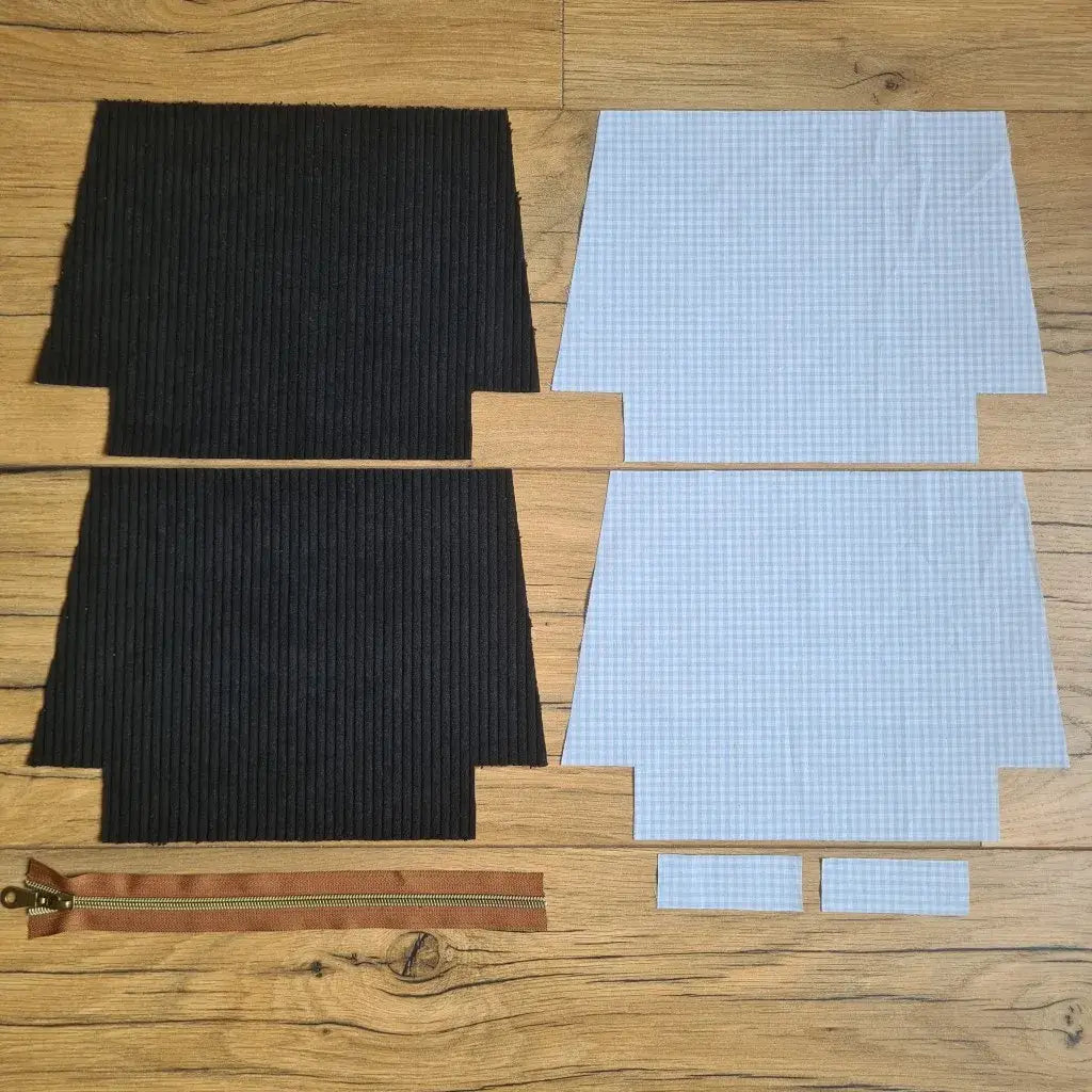 Kit "prêt à coudre" pour une trousse zippée en velours côtelé au coloris noir avec une doublure en tissu à motif carreaux bleus. L'ensemble des pièces de tissu, déjà prédécoupées aux bonnes dimensions, sont disposées au sol. A gauche, deux pièces identiques en velours côtelé noir ainsi qu'une fermeture éclair marron. A droite, quatre pièces en tissu imprimé à motif carreaux bleus.