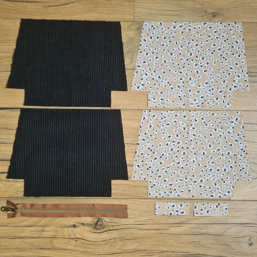 Kit "prêt à coudre" pour une trousse zippée en velours côtelé au coloris noir avec une doublure en tissu à motif léopard. L'ensemble des pièces de tissu, déjà prédécoupées aux bonnes dimensions, sont disposées au sol. A gauche, deux pièces identiques en velours côtelé noir ainsi qu'une fermeture éclair marron. A droite, quatre pièces en tissu imprimé à motif léopard.