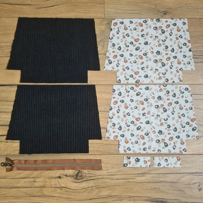 Kit "prêt à coudre" pour une trousse zippée en velours côtelé au coloris noir avec une doublure en tissu à motif léopard. L'ensemble des pièces de tissu, déjà prédécoupées aux bonnes dimensions, sont disposées au sol. A gauche, deux pièces identiques en velours côtelé noir ainsi qu'une fermeture éclair marron. A droite, quatre pièces en tissu imprimé à motif léopard.