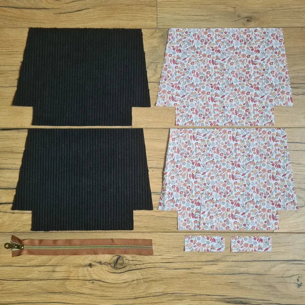 Kit "prêt à coudre" pour une trousse zippée en velours côtelé au coloris noir avec une doublure en tissu à motif liberty. L'ensemble des pièces de tissu, déjà prédécoupées aux bonnes dimensions, sont disposées au sol. A gauche, deux pièces identiques en velours côtelé noir ainsi qu'une fermeture éclair marron. A droite, quatre pièces en tissu imprimé à motif liberty.