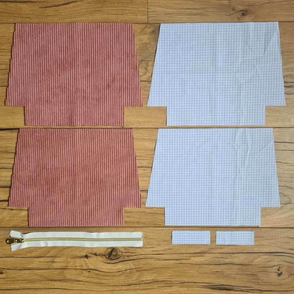 Kit "prêt à coudre" pour une trousse zippée en velours côtelé au coloris rose avec une doublure en tissu à motif carreaux bleus. L'ensemble des pièces de tissu, déjà prédécoupées aux bonnes dimensions, sont disposées au sol. A gauche, deux pièces identiques en velours côtelé rose ainsi qu'une fermeture éclair blanche. A droite, quatre pièces en tissu imprimé à motif carreaux bleus.