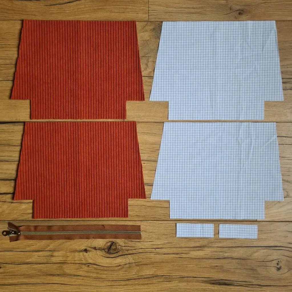 Kit "prêt à coudre" pour une trousse zippée en velours côtelé au coloris terracotta avec une doublure en tissu à motif carreaux bleus. L'ensemble des pièces de tissu, déjà prédécoupées aux bonnes dimensions, sont disposées au sol. A gauche, deux pièces identiques en velours côtelé terracotta ainsi qu'une fermeture éclair marron. A droite, quatre pièces en tissu imprimé à motif carreaux bleus.