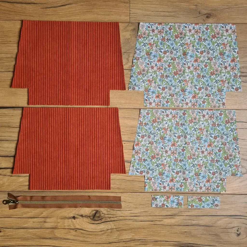 Kit "prêt à coudre" pour une trousse zippée en velours côtelé au coloris terracotta avec une doublure en tissu à motif fleuri. L'ensemble des pièces de tissu, déjà prédécoupées aux bonnes dimensions, sont disposées au sol. A gauche, deux pièces identiques en velours côtelé terracotta ainsi qu'une fermeture éclair marron. A droite, quatre pièces en tissu imprimé à motif fleuri.