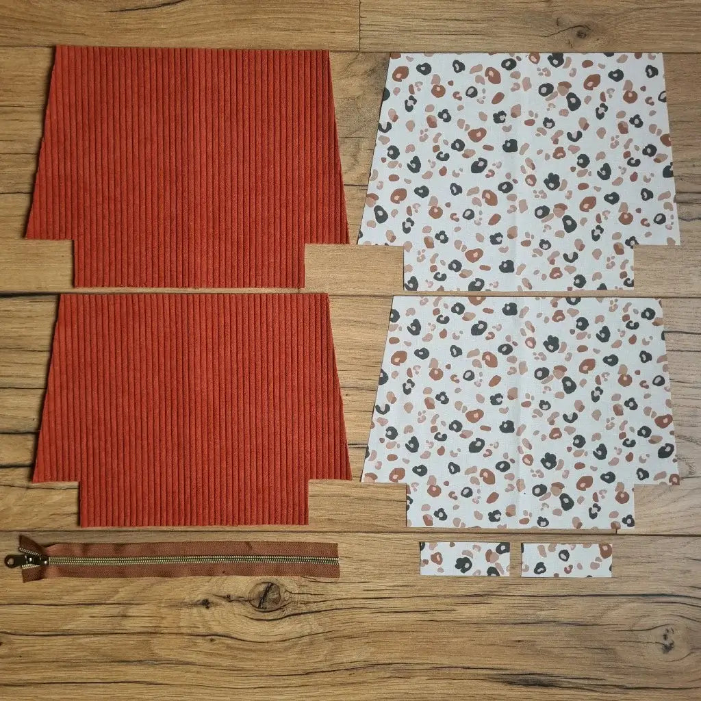 Kit "prêt à coudre" pour une trousse zippée en velours côtelé au coloris terracotta avec une doublure en tissu à motif léopard. L'ensemble des pièces de tissu, déjà prédécoupées aux bonnes dimensions, sont disposées au sol. A gauche, deux pièces identiques en velours côtelé terracotta ainsi qu'une fermeture éclair marron. A droite, quatre pièces en tissu imprimé à motif léopard.