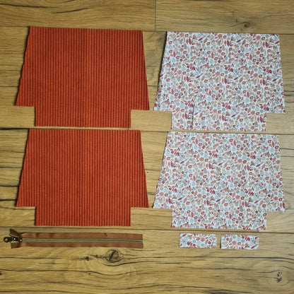 Kit "prêt à coudre" pour une trousse zippée en velours côtelé au coloris terracotta avec une doublure en tissu à motif liberty. L'ensemble des pièces de tissu, déjà prédécoupées aux bonnes dimensions, sont disposées au sol. A gauche, deux pièces identiques en velours côtelé terracotta ainsi qu'une fermeture éclair marron. A droite, quatre pièces en tissu imprimé à motif liberty.