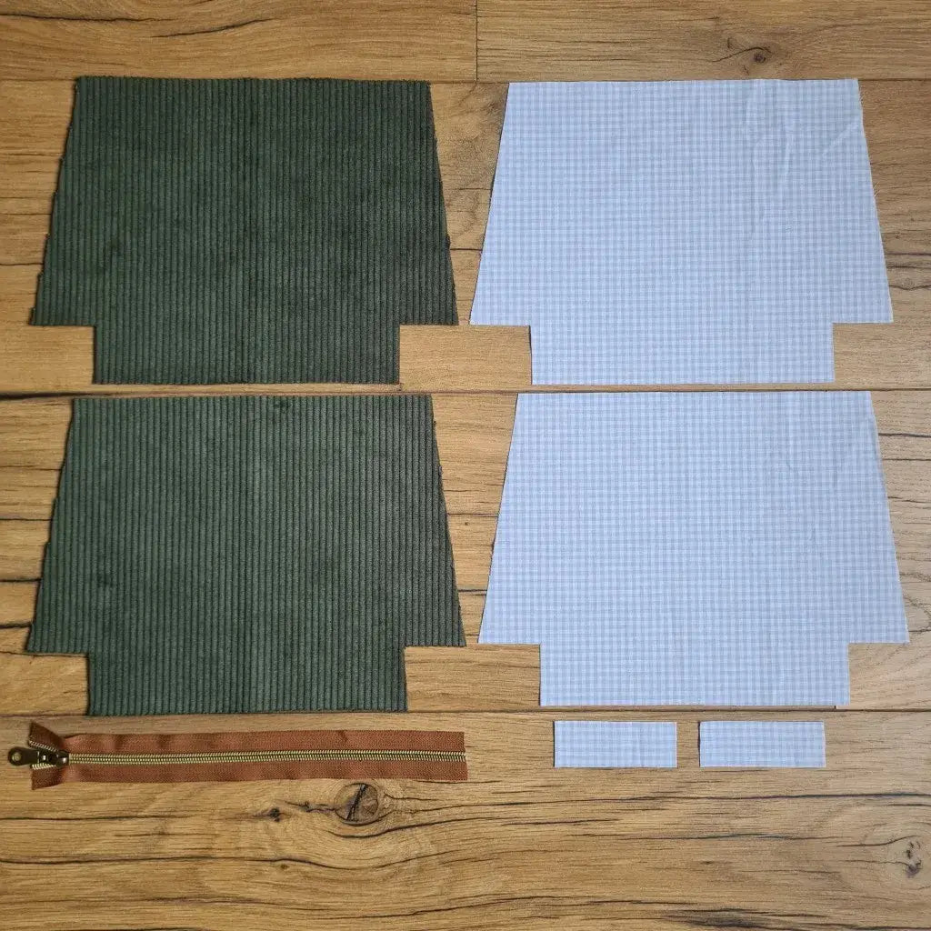 Kit "prêt à coudre" pour une trousse zippée en velours côtelé au coloris vert avec une doublure en tissu à motif carreaux bleus. L'ensemble des pièces de tissu, déjà prédécoupées aux bonnes dimensions, sont disposées au sol. A gauche, deux pièces identiques en velours côtelé vert ainsi qu'une fermeture éclair marron. A droite, quatre pièces en tissu imprimé à motif carreaux bleus.