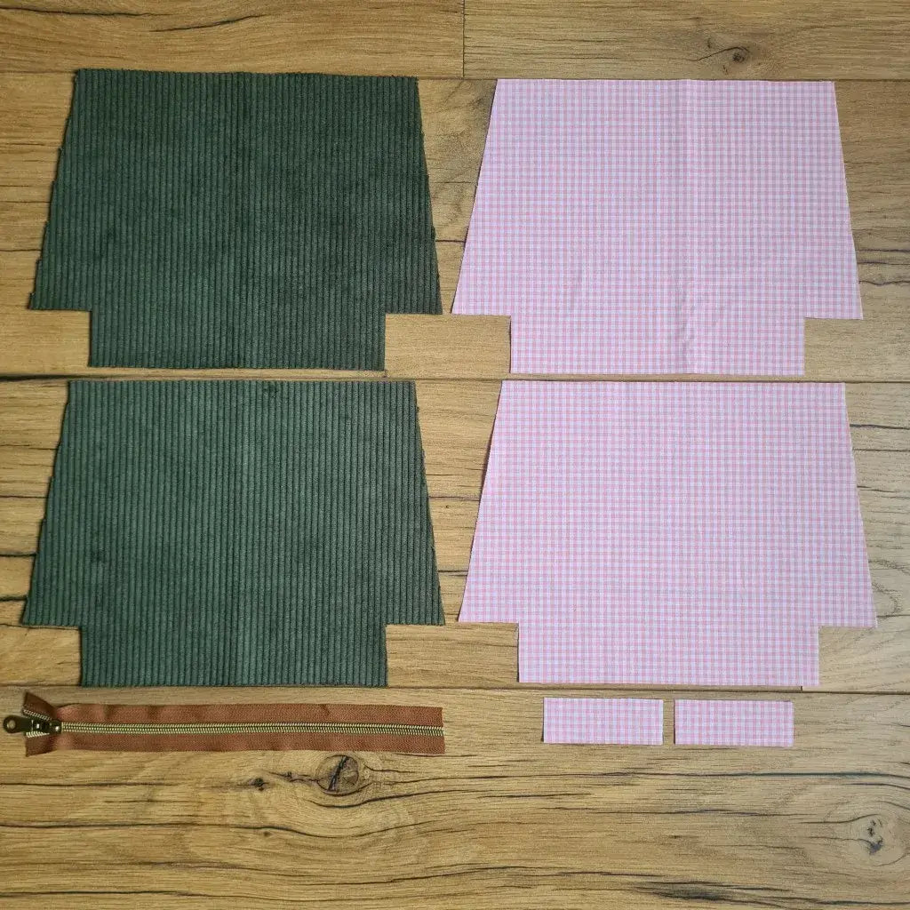 Kit "prêt à coudre" pour une trousse zippée en velours côtelé au coloris vert avec une doublure en tissu à motif carreaux roses. L'ensemble des pièces de tissu, déjà prédécoupées aux bonnes dimensions, sont disposées au sol. A gauche, deux pièces identiques en velours côtelé vert ainsi qu'une fermeture éclair marron. A droite, quatre pièces en tissu imprimé à motif carreaux roses.