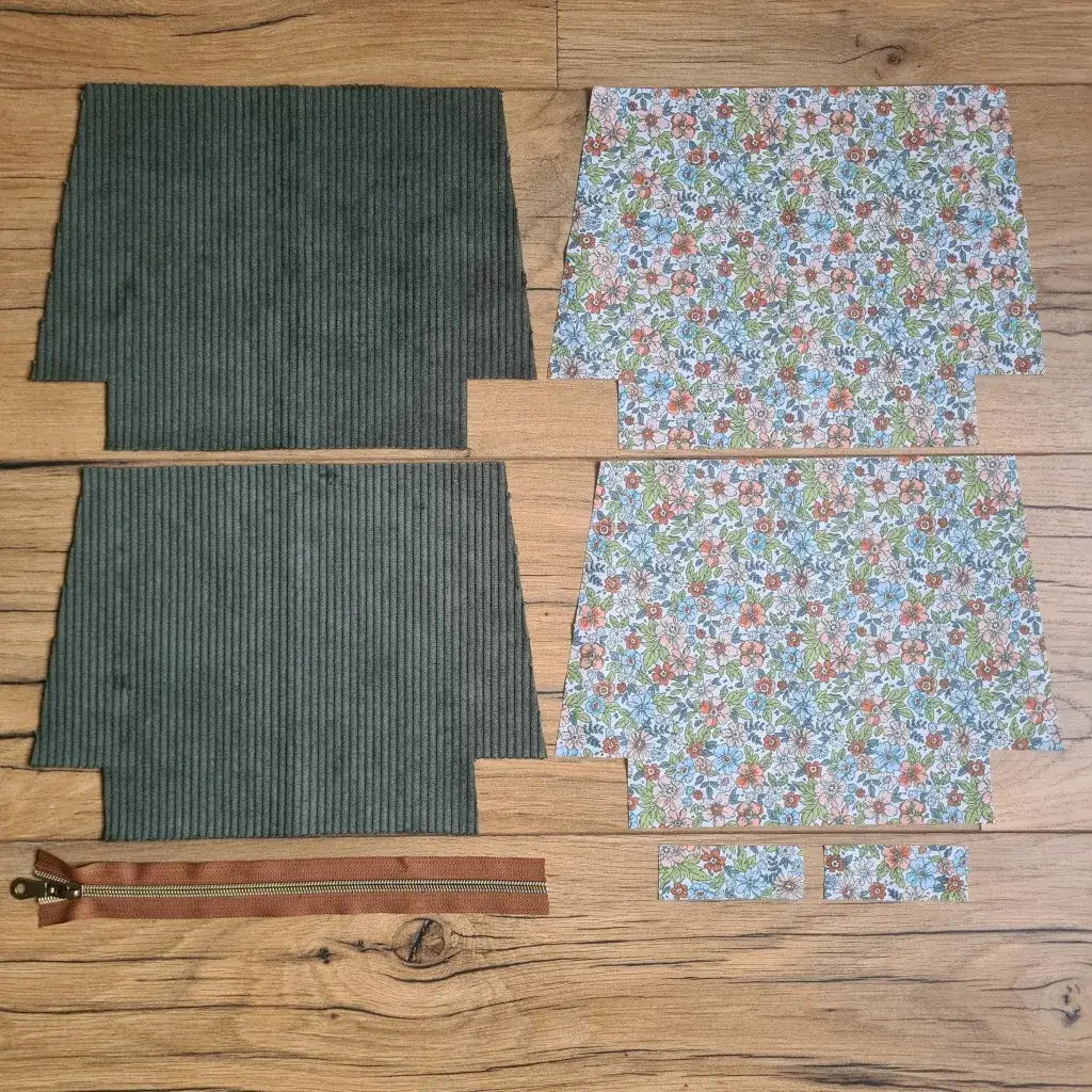 Kit "prêt à coudre" pour une trousse zippée en velours côtelé au coloris vert avec une doublure en tissu à motif fleuri. L'ensemble des pièces de tissu, déjà prédécoupées aux bonnes dimensions, sont disposées au sol. A gauche, deux pièces identiques en velours côtelé vert ainsi qu'une fermeture éclair marron. A droite, quatre pièces en tissu imprimé à motif fleuri.