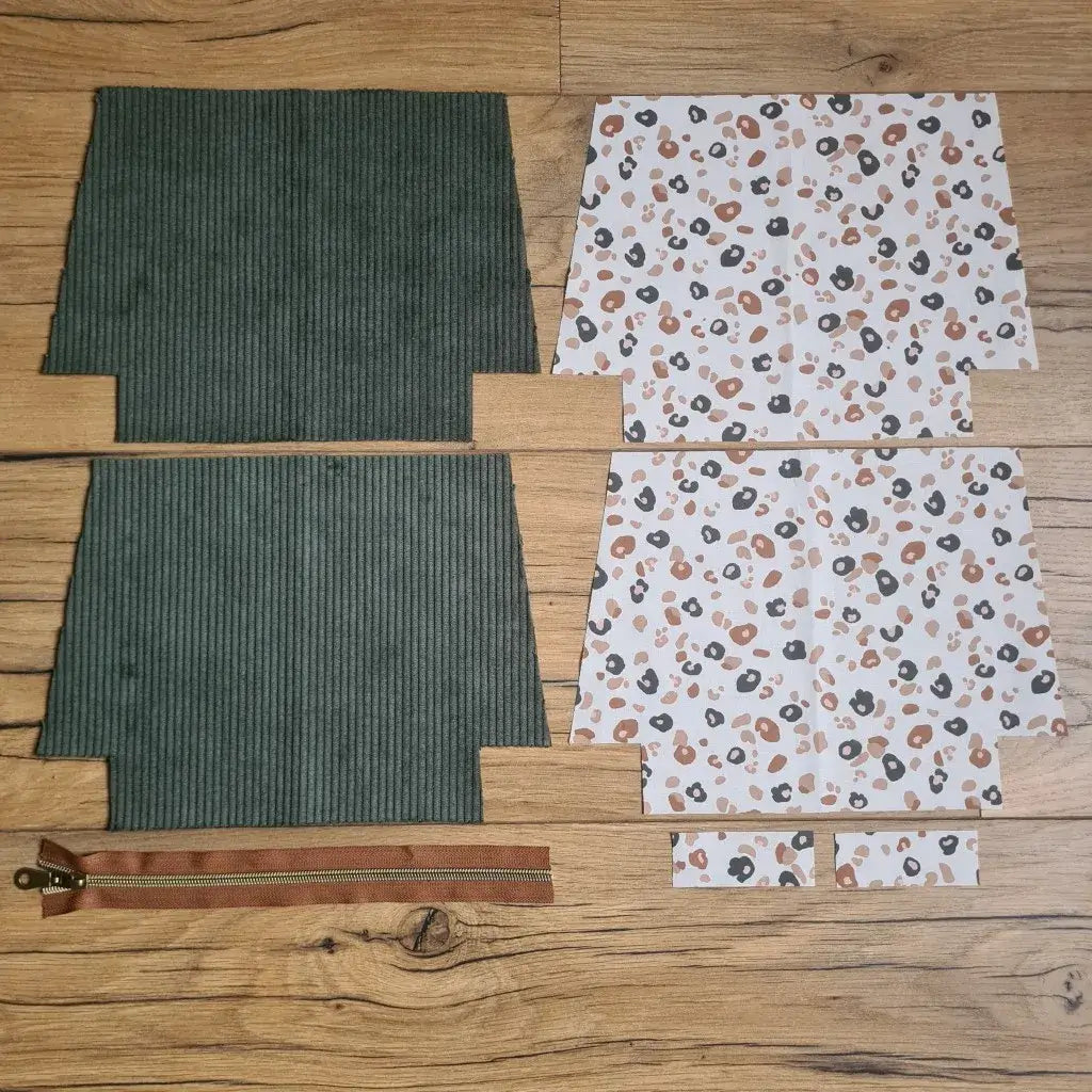 Kit "prêt à coudre" pour une trousse zippée en velours côtelé au coloris vert avec une doublure en tissu à motif léopard. L'ensemble des pièces de tissu, déjà prédécoupées aux bonnes dimensions, sont disposées au sol. A gauche, deux pièces identiques en velours côtelé vert ainsi qu'une fermeture éclair marron. A droite, quatre pièces en tissu imprimé à motif léopard.