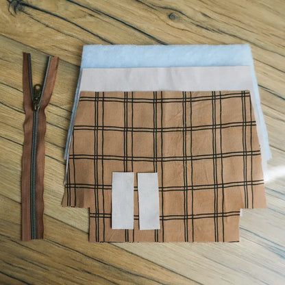 Kit couture complet pour confectionner une trousse zippée en tissu à carreaux marron et noir. L’ensemble comprend une fermeture éclair marron avec curseur métallique, des pièces de tissu découpées, une doublure unie claire et de la ouate pour plus de tenue. Parfait pour apprendre la couture et créer une trousse fait main pratique et personnalisée.
