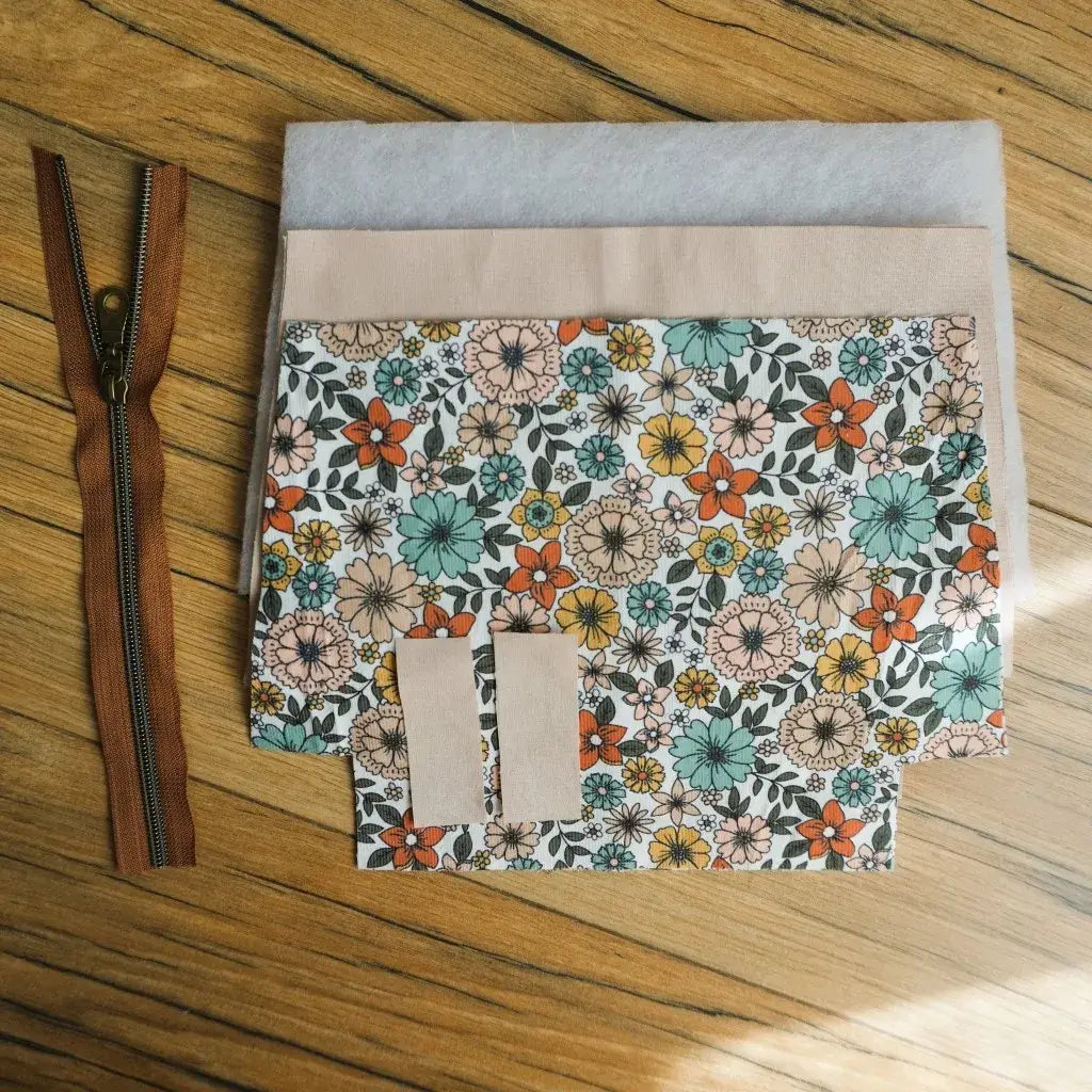 Kit de couture DIY pour créer une trousse zippée en tissu fleuri rétro aux tons orange, jaune, vert et rose. Le set contient une fermeture éclair marron avec curseur métallique, des pièces de tissu prédécoupées, une doublure unie claire et de la ouate pour assurer maintien et solidité. Un projet couture facile et créatif pour fabriquer une trousse fait main pratique et personnalisée.