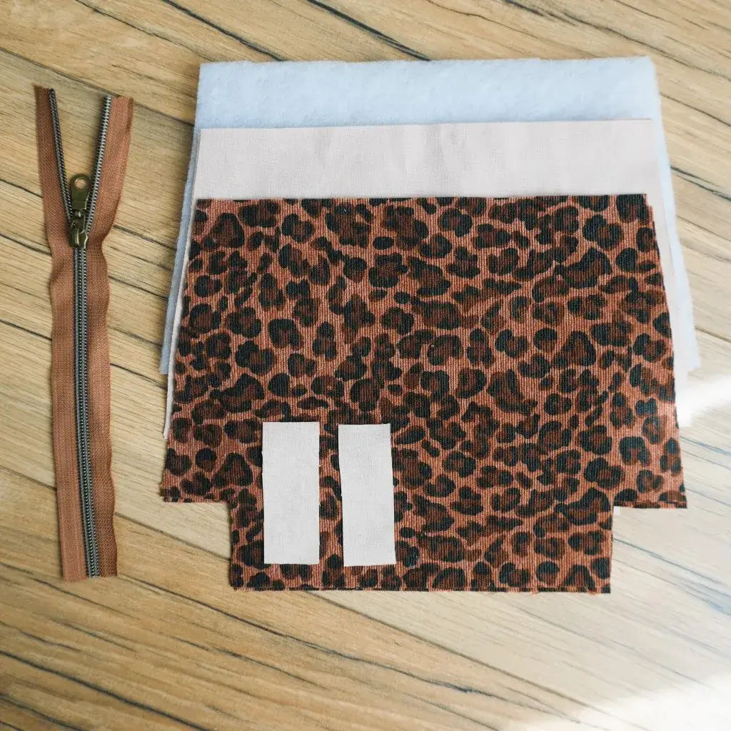 Kit couture DIY pour confectionner une trousse zippée en tissu velours côtelé motif léopard marron et noir. Le kit comprend une fermeture éclair marron avec curseur métallique, des pièces de tissu prédécoupées, une doublure claire et de la ouate pour assurer solidité et tenue. Idéal pour un projet créatif fait main, permettant de réaliser une trousse pratique et personnalisée au style tendance.