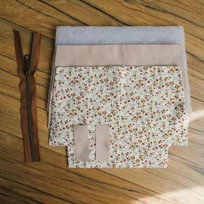 Kit prêt-à-coudre pour réaliser une trousse zippée en tissu imprimé fleuri style prairie, aux petites fleurs orange et jaunes sur fond clair. Le kit contient une fermeture éclair marron avec curseur métallique, des morceaux de tissu découpés, une doublure beige et deux pièces de ouate pour renforcer la tenue. Un projet DIY couture idéal pour créer une trousse fait main pratique et personnalisée.
