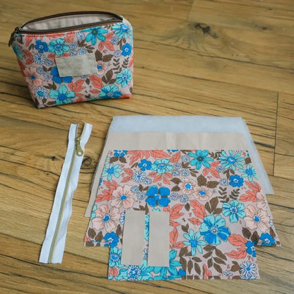 Kit prêt-à-coudre pour réaliser une trousse zippée en tissu fleuri aux tons corail, bleu et marron. L’image montre la trousse terminée avec doublure beige et étiquette en tissu, ainsi que les fournitures nécessaires : fermeture éclair blanche avec curseur doré, pièces de tissu découpées, doublure et ouate. Idéal pour la couture créative et les accessoires faits main personnalisés.