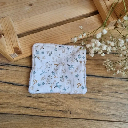 Lingette démaquillante lavable en tissu fleuri aux tons bleus et blancs, posée sur un sol en bois. Accessoire écologique, réutilisable et zéro déchet, idéal pour une routine beauté respectueuse de l’environnement. Motif floral délicat et minimaliste, parfait pour une touche de douceur et d’élégance. Fabrication artisanale avec couture soignée et texture ultra-douce en coton et éponge de bambou, garantissant un nettoyage du visage tout en confort.