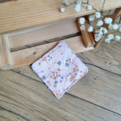 Lingette démaquillante lavable en tissu fleuri posée sur un sol en bois. Accessoire zéro déchet, réutilisable et écologique, idéal pour une routine beauté durable. Motif floral délicat sur fond rose, couture soignée et finition douce. Parfait pour un nettoyage du visage tout en douceur. Fabrication artisanale et respectueuse de l’environnement.