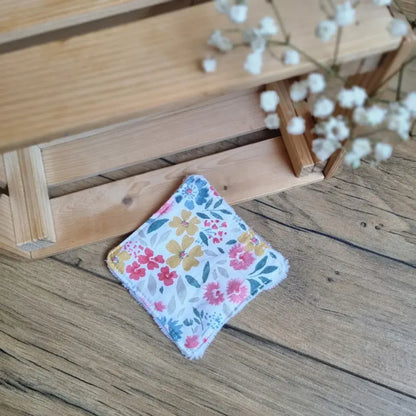 Lingette démaquillante lavable en tissu aux motifs floraux colorés, posée sur un sol en bois. Accessoire de beauté écologique, zéro déchet et réutilisable, idéal pour une routine durable. Motifs fleuris aux teintes vives – jaune, rose et bleu – apportant une touche de fraîcheur et de douceur. Confection artisanale avec des finitions soignées pour un nettoyage du visage tout en respectant la peau et l’environnement.