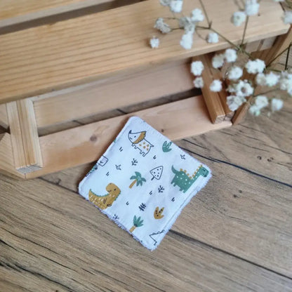 Lingette démaquillante lavable en tissu blanc avec motifs dinosaures, posée sur un sol en bois. Accessoire réutilisable, écologique et zéro déchet, idéal pour une routine beauté durable. Imprimé ludique avec dinosaures, palmiers et petits éléments graphiques, parfait pour les amateurs de motifs enfantins et originaux. Fabrication artisanale avec des finitions soignées et un toucher doux, respectueux de la peau et de l’environnement.
