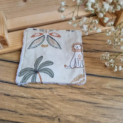 Lingette démaquillante lavable en tissu motif jungle, posée sur un sol en bois. Accessoire réutilisable, zéro déchet et écologique, parfait pour une routine beauté responsable. Imprimé inspiré de la savane avec lions, girafes, zèbres et palmiers, apportant une touche exotique et apaisante. Fabrication artisanale avec couture soignée et texture ultra-douce en coton et éponge de bambou, idéale pour un nettoyage du visage tout en confort et respect de l’environnement.