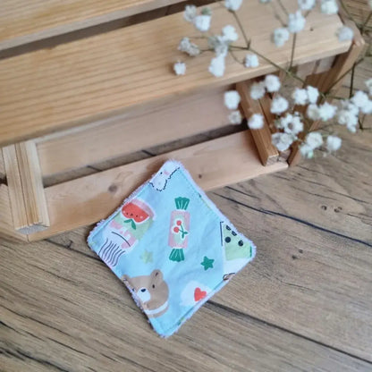 Lingette démaquillante lavable en tissu bleu pastel avec motifs ludiques d’animaux et de gourmandises, posée sur un sol en bois. Accessoire de beauté réutilisable, écologique et zéro déchet, idéal pour une routine de soin respectueuse de l’environnement. Imprimé kawaii avec ourson, pastèques, bonbons et étoiles, apportant une touche joyeuse et enfantine. Fabrication artisanale, couture soignée et texture douce pour un nettoyage du visage tout en délicatesse.
