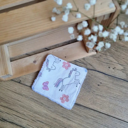 Lingette démaquillante lavable en tissu blanc avec motifs licornes, posée sur un sol en bois. Accessoire réutilisable, écologique et zéro déchet, idéal pour une routine beauté durable. Imprimé féerique avec licornes, parfait pour une touche de magie et de tendresse. Fabrication artisanale, couture soignée et texture ultra-douce pour un nettoyage du visage tout en confort et respect de l’environnement.