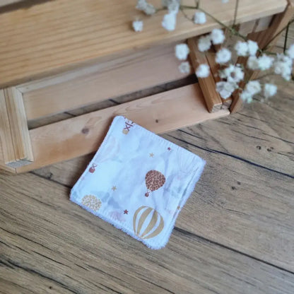 Lingette démaquillante lavable en tissu blanc avec motifs de montgolfières dorées et marron, posée sur un sol en bois. Accessoire écologique, réutilisable et zéro déchet, parfait pour une routine beauté responsable. Imprimé avec montgolfières, idéal pour une touche de légèreté et d’évasion. Fabrication artisanale, couture soignée et texture ultra-douce pour un nettoyage du visage respectueux de la peau et de l’environnement.
