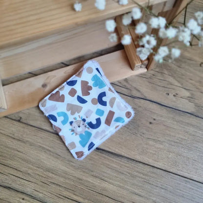 Lingette démaquillante lavable en tissu aux motifs géométriques bleus et marron, posée sur un sol en bois. Accessoire réutilisable, zéro déchet et écologique, idéal pour une routine beauté durable. Design moderne, couture soignée et toucher doux. Parfaite pour un nettoyage du visage tout en douceur. Fabrication artisanale et respectueuse de l’environnement.