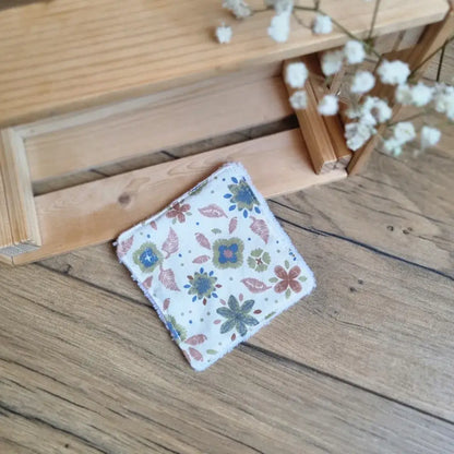 Lingette démaquillante lavable en tissu fleuri aux tons verts et marron, posée sur un sol en bois. Accessoire écologique, zéro déchet et réutilisable, parfait pour une routine beauté durable. Motifs floraux délicats inspirés de la nature, couture soignée et douceur au toucher. Idéale pour le nettoyage du visage tout en respectant la peau et l’environnement. Fabrication artisanale et responsable.