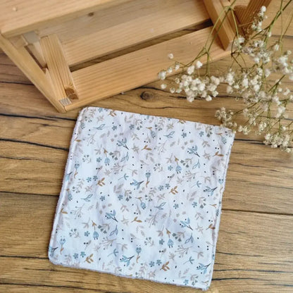 Lingette démaquillante lavable au format maxi 18x18 cm en tissu fleuri aux tons bleus et blancs, posée sur un sol en bois. Accessoire écologique, réutilisable et zéro déchet, idéal pour une routine beauté respectueuse de l’environnement. Motif floral délicat et minimaliste, parfait pour une touche de douceur et d’élégance. Fabrication artisanale avec couture soignée et texture ultra-douce en coton et éponge de bambou, garantissant un nettoyage du visage tout en confort.
