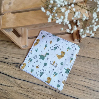 Lingette démaquillante lavable au format maxi 18x18 cm en tissu blanc avec motifs dinosaures, posée sur un sol en bois. Accessoire réutilisable, écologique et zéro déchet, idéal pour une routine beauté durable. Imprimé ludique avec dinosaures, palmiers et petits éléments graphiques, parfait pour les amateurs de motifs enfantins et originaux. Fabrication artisanale avec des finitions soignées et un toucher doux, respectueux de la peau et de l’environnement.