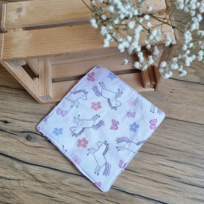 Lingette démaquillante lavable au format maxi 18x18 cm en tissu blanc avec motifs licornes, posée sur un sol en bois. Accessoire réutilisable, écologique et zéro déchet, idéal pour une routine beauté durable. Imprimé féerique avec licornes, parfait pour une touche de magie et de tendresse. Fabrication artisanale, couture soignée et texture ultra-douce pour un nettoyage du visage tout en confort et respect de l’environnement.