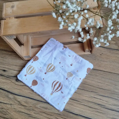 Lingette démaquillante lavable au format maxi 18x18 cm en tissu blanc avec motifs de montgolfières dorées et marron, posée sur un sol en bois. Accessoire écologique, réutilisable et zéro déchet, parfait pour une routine beauté responsable. Imprimé avec montgolfières, idéal pour une touche de légèreté et d’évasion. Fabrication artisanale, couture soignée et texture ultra-douce pour un nettoyage du visage respectueux de la peau et de l’environnement.