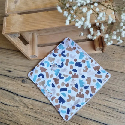 Lingette démaquillante lavable format maxi 18x18 cm en tissu aux motifs géométriques bleus et marron, posée sur un sol en bois. Accessoire réutilisable, zéro déchet et écologique, idéal pour une routine beauté durable. Design moderne, couture soignée et toucher doux. Parfaite pour un nettoyage du visage tout en douceur. Fabrication artisanale et respectueuse de l’environnement.