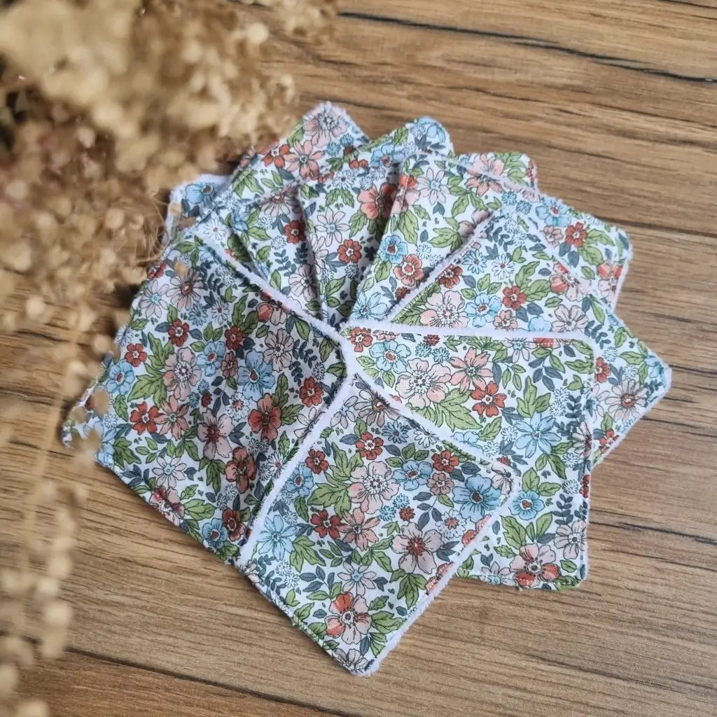 Tas de 10 lingettes en coton lavables et réutilisables au format classique 10x10 cm disposées en éventail sur un parquet en chêne. Ces lingettes présentent une face en coton imprimé d'un motif fleuri.