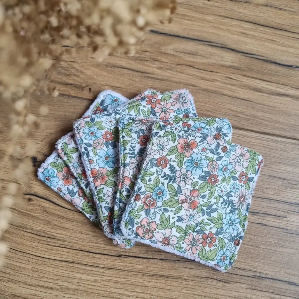 Tas de 5 lingettes en coton lavables et réutilisables au format classique 10x10 cm disposées en éventail sur un parquet en chêne. Ces lingettes présentent une face en coton imprimé d'un motif fleuri.