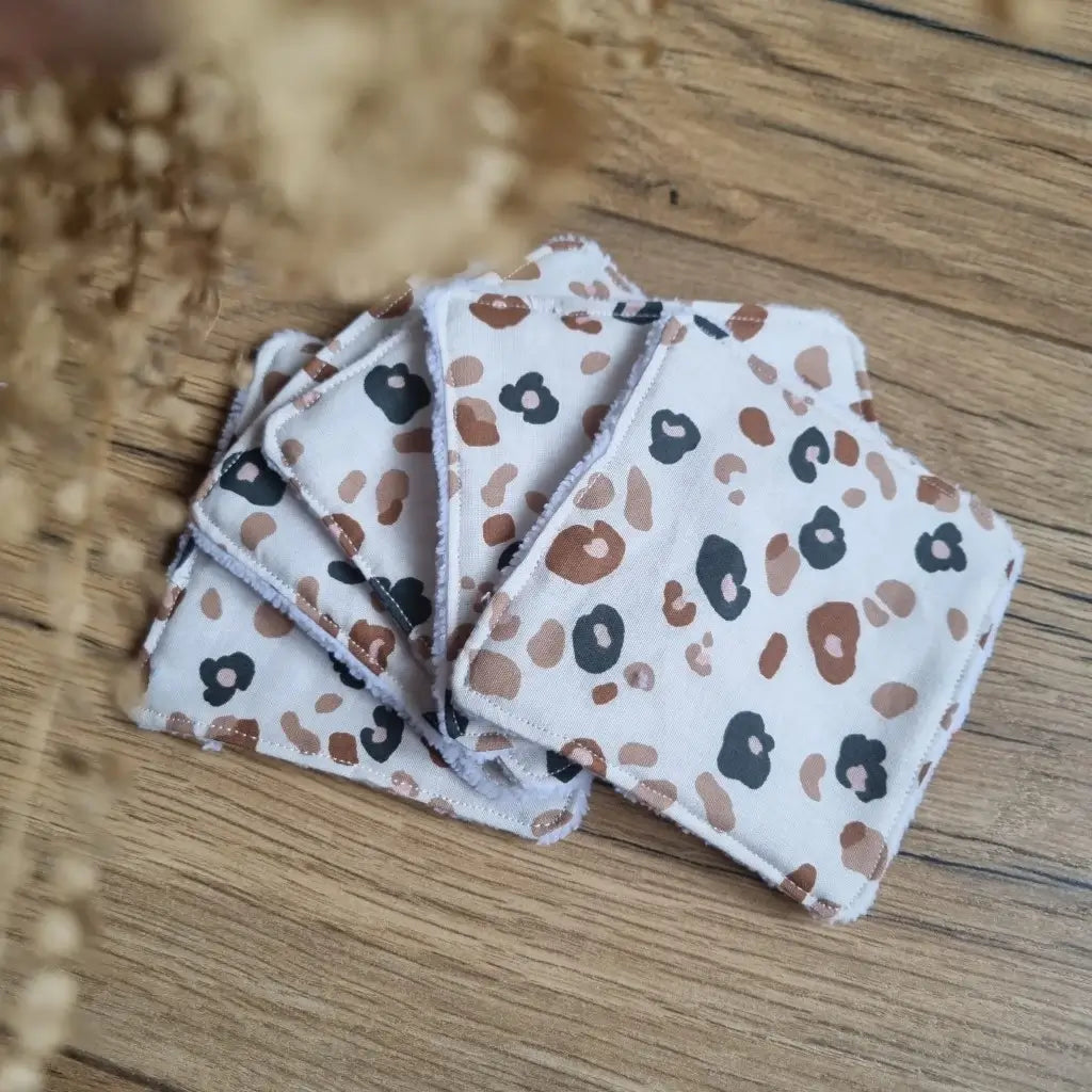 Tas de 5 lingettes en coton lavables et réutilisables au format classique 10x10 cm disposées en éventail sur un parquet en chêne. Ces lingettes présentent une face en coton imprimé d'un motif léopard.