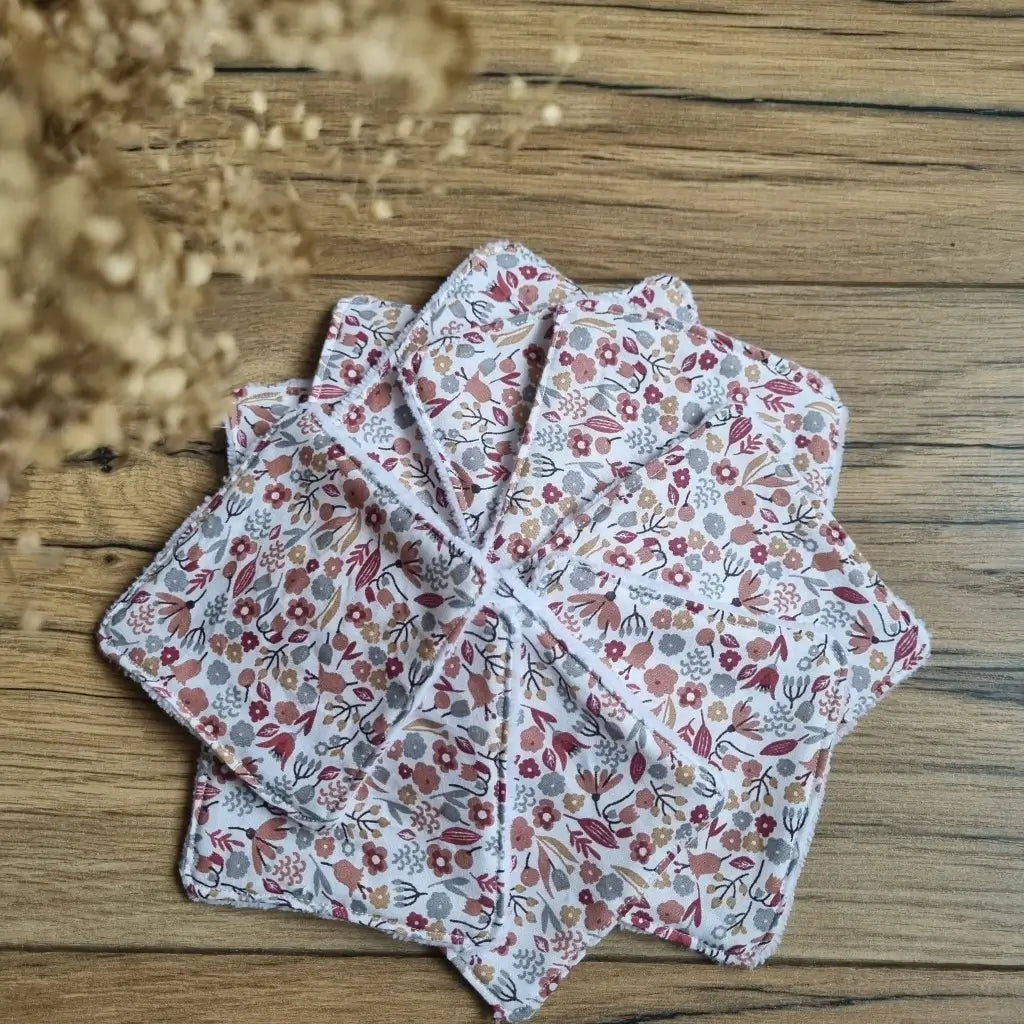 Tas de 10 lingettes en coton lavables et réutilisables au format classique 10x10 cm disposées en éventail sur un parquet en chêne. Ces lingettes présentent une face en coton imprimé d'un motif liberty.