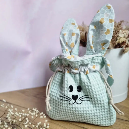 Panier de Pâques en tissu en forme de lapin, confectionné en coton nid d’abeille couleur menthe avec des oreilles en tissu imprimé. Ce pochon réutilisable avec lien de serrage en corde naturelle est idéal pour la chasse aux œufs ou comme emballage cadeau écologique. Son design ludique avec un visage de lapin floqué apporte une touche festive et originale à Pâques. Accessoire pratique et durable pour petits et grands.