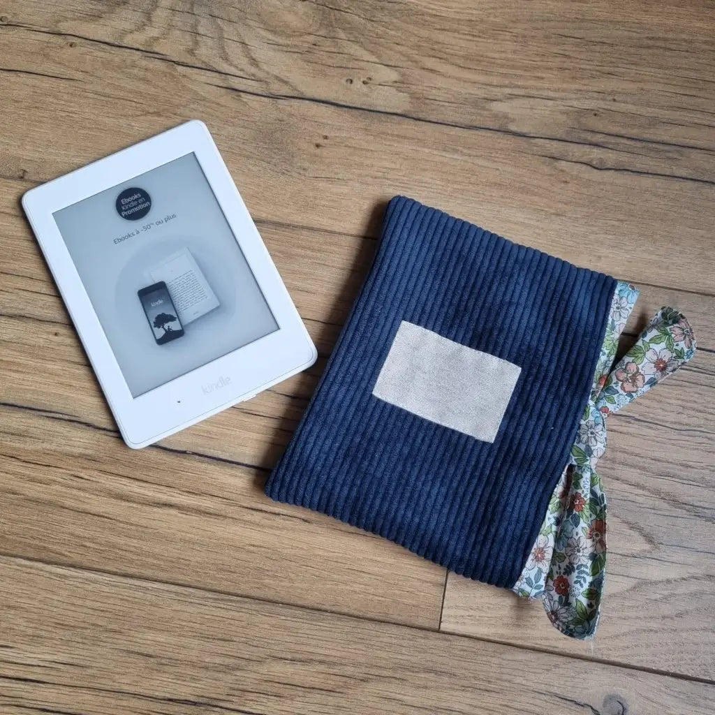 A droite, une pochette à livre ou liseuse en velours côtelé bleu foncé fermée à l'aide d'un ruban en tissu imprimé fleuri. Cette pochette comporte une étiquette cousue en son centre sans inscription. A gauche, une liseuse type Kindle de coloris blanc.