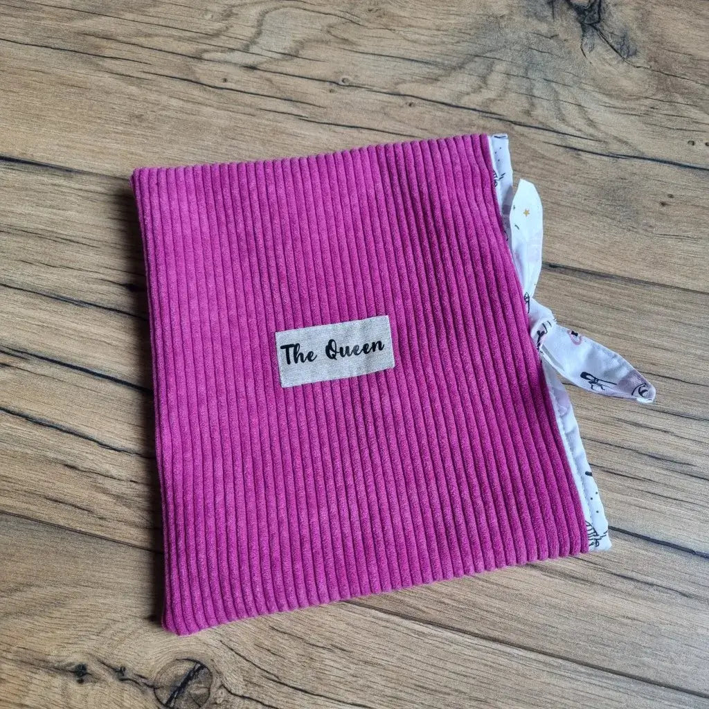 Pochette à livre ou étui pour liseuse en velours côtelé magenta, doublée d’un tissu imprimé blanc à motifs, fermée par un joli nœud latéral. Accessoire de lecture chic et doux au toucher, idéal pour protéger un roman ou une tablette en déplacement. Étiquette “The Queen” cousue sur le devant pour une touche élégante et personnalisée.