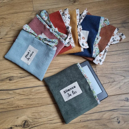 Au premier plan, une pochette à livre ou liseuse en velours côtelé vert avec une étiquette cousue en son centre portant l'inscription "Silence Je lis". Au-dessus de celle-ci, six autres pochettes semblables disposées en éventail et de coloris divers (de gauche à droite : bleu ciel, rose, terracotta, jaune, bleu foncé et marron).