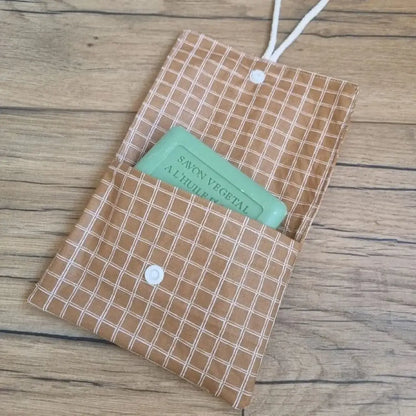 Pochette à savon double compartiment en coton enduit au motif carreaux. Cette pochette dispose d'une petite cordelette en coton blanc lui permettant d'être suspendue dans la douche. Un savon vert à l'huile d'olive est glissé à moitié à l'intérieur de cette pochette.