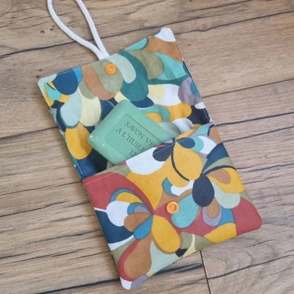 Pochette à savon double compartiment en coton enduit au motif splash. Cette pochette dispose d'une petite cordelette en coton blanc lui permettant d'être suspendue dans la douche. Un savon vert à l'huile d'olive est glissé à moitié à l'intérieur de cette pochette.