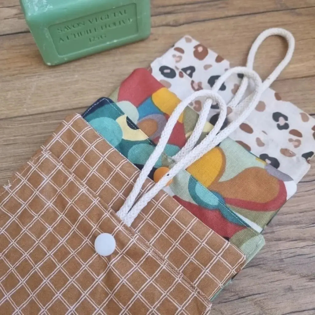 Trois pochettes à savon double compartiment en coton enduit de divers motifs sont alignés les uns par-dessus les autres. Ces pochettes disposent d'une cordelette en coton blanc leur permettant d'être suspendues à l'intérieur de la douche. A l'arrière plan, on distingue un savon vert à l'huile d'olive posé juste à côté des pochettes à savon.