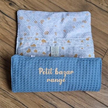 Pochette de rangement multi-usages en nid d'abeille de coloris bleu denim, doublée avec un tissu imprimé à motifs Argeo et personnalisée avec l'inscription "Petit bazar rangé" floqué en doré. Cette pochette est à moitié enroulée sur elle-même.