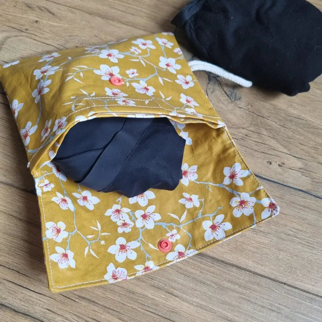 Pochette double compartiment en coton enduit au motif fleurs jaunes pour culottes menstruelles ou serviettes hygiéniques lavables. La pochette est ouverte et laisse apparaître une culotte menstruelle noire rangée à l'intérieur. A l'extérieur, une seconde culotte menstruelle noire est roulée et disposée à côté de la pochette.