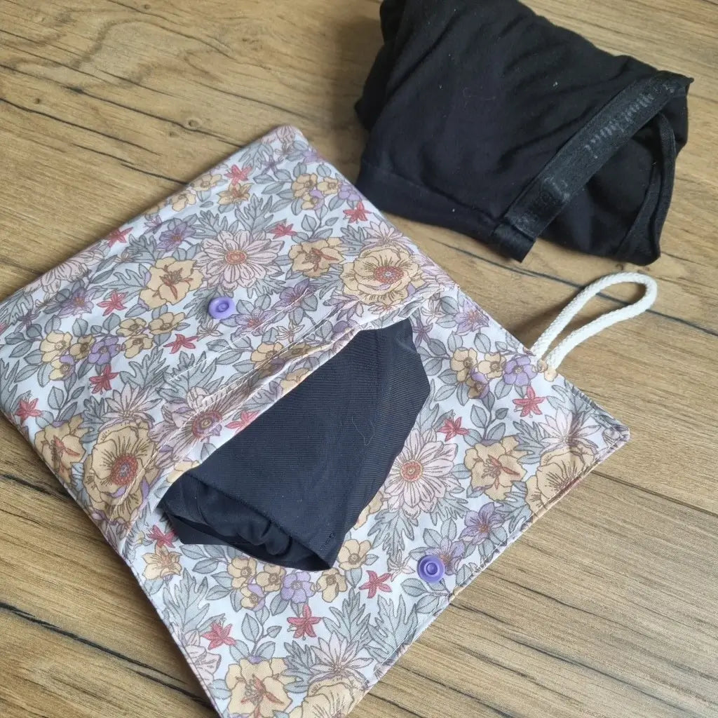 Pochette double compartiment en coton enduit au motif fleurs mauves pour culottes menstruelles ou serviettes hygiéniques lavables. La pochette est ouverte et laisse apparaître une culotte menstruelle noire rangée à l'intérieur. A l'extérieur, une seconde culotte menstruelle noire est roulée et disposée à côté de la pochette.