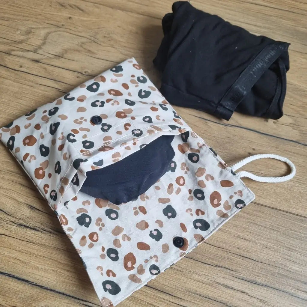 Pochette double compartiment en coton enduit au motif léopard pour culottes menstruelles ou serviettes hygiéniques lavables. La pochette est ouverte et laisse apparaître une culotte menstruelle noire rangée à l'intérieur. A l'extérieur, une seconde culotte menstruelle noire est roulée et disposée à côté de la pochette.