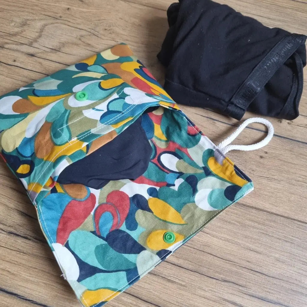 Pochette double compartiment en coton enduit au motif splash pour culottes menstruelles ou serviettes hygiéniques lavables. La pochette est ouverte et laisse apparaître une culotte menstruelle noire rangée à l'intérieur. A l'extérieur, une seconde culotte menstruelle noire est roulée et disposée à côté de la pochette.