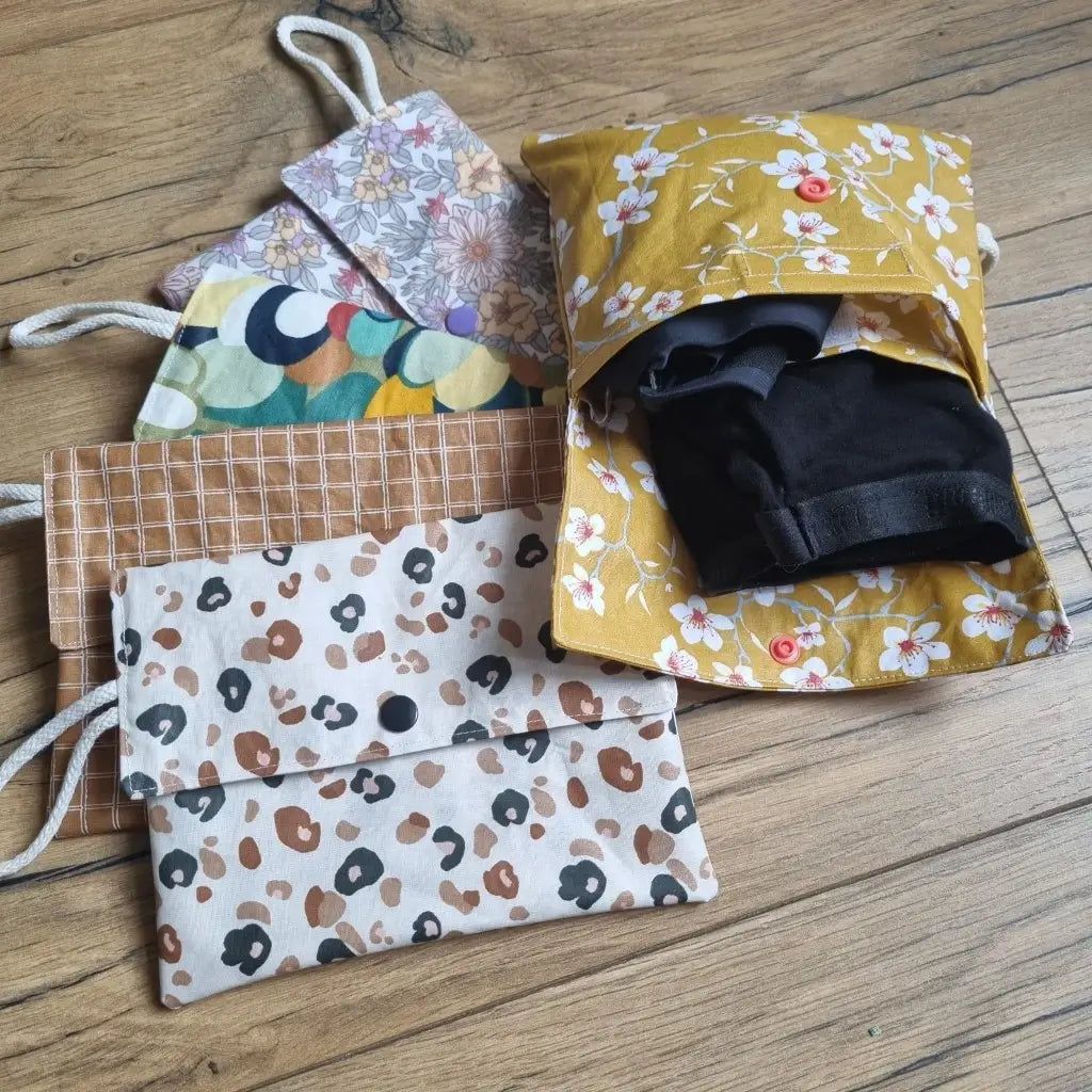 Pochettes double compartiment en coton enduit pour culottes menstruelles ou serviettes hygiéniques lavables. La pochette de droite, au motif fleurs jaunes, est ouverte et deux culottes menstruelles sont rangées à l'intérieur. A gauche sont disposées en éventail quatre autres pochettes fermées. Les coloris de haut en bas sont les suivants : fleurs mauves, splash, carreaux et léopard.